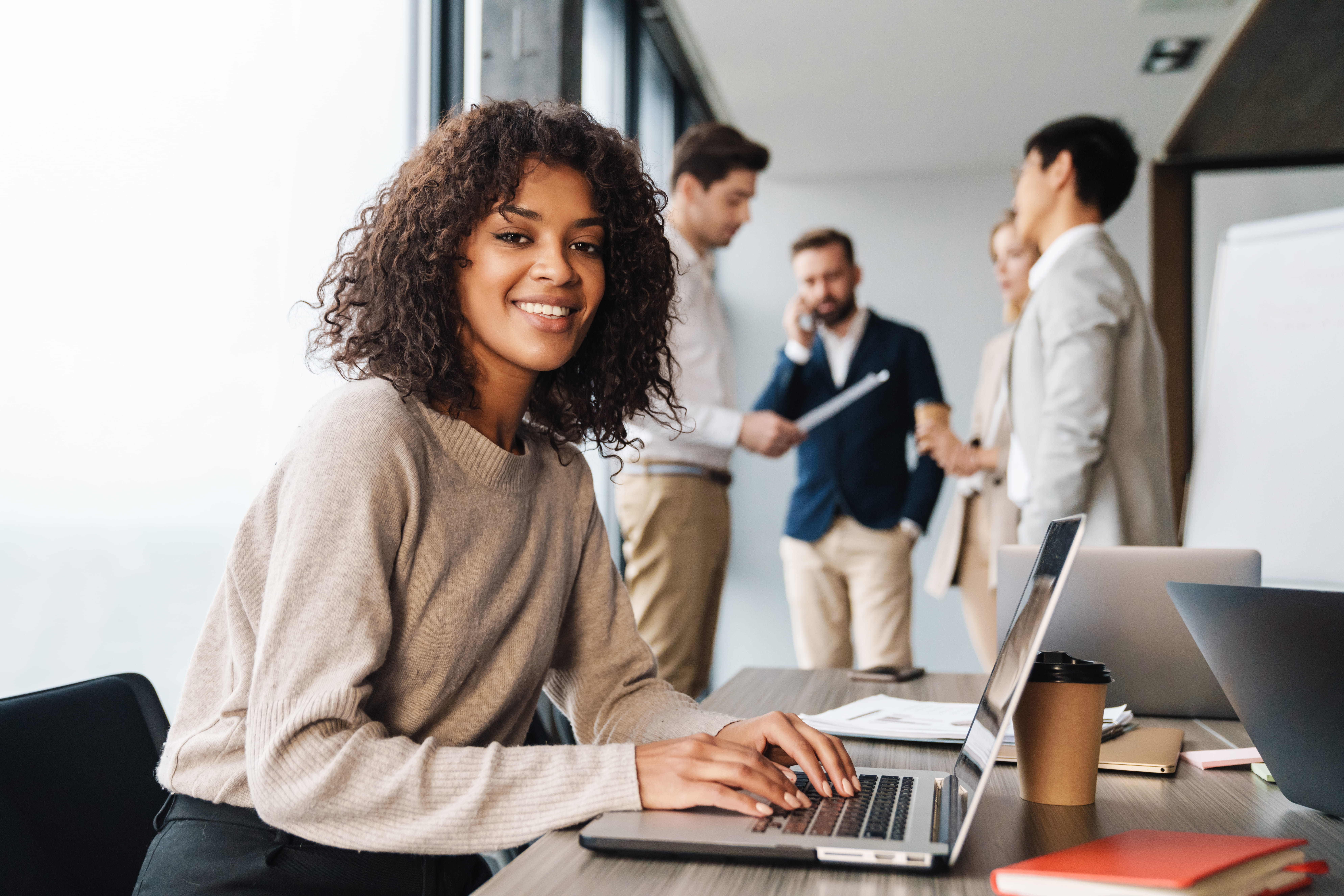 Smiling person working on a laptop seated at desk with coworkers in the background.