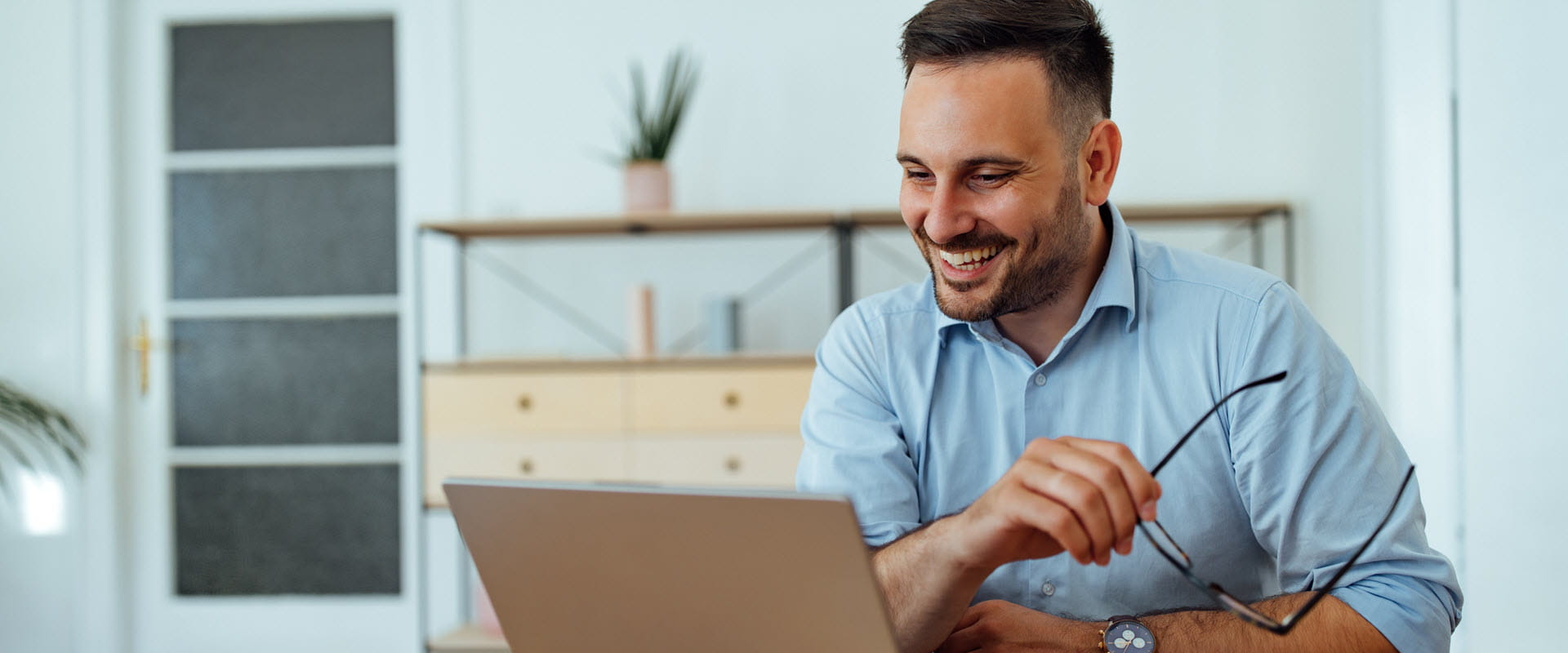 A smiling individual sitting at a table, engaging in a virtual meeting on a laptop while holding glasses in one hand. 